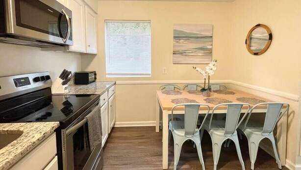 A compact kitchen with a microwave, oven, and toaster. There is a dining table with four chairs and a vase with flowers. - ★Pristine Pad ★ King Bed ★Duke ★ Long Term Stays★ (Durham)