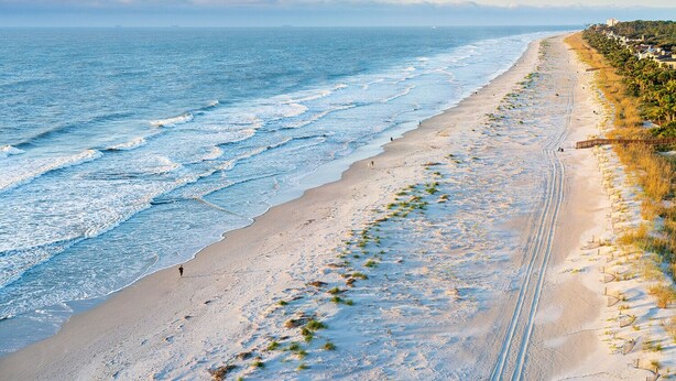 Una playa con vista despejada al océano, un sendero arenoso y una hilera de casas a lo largo de la costa.