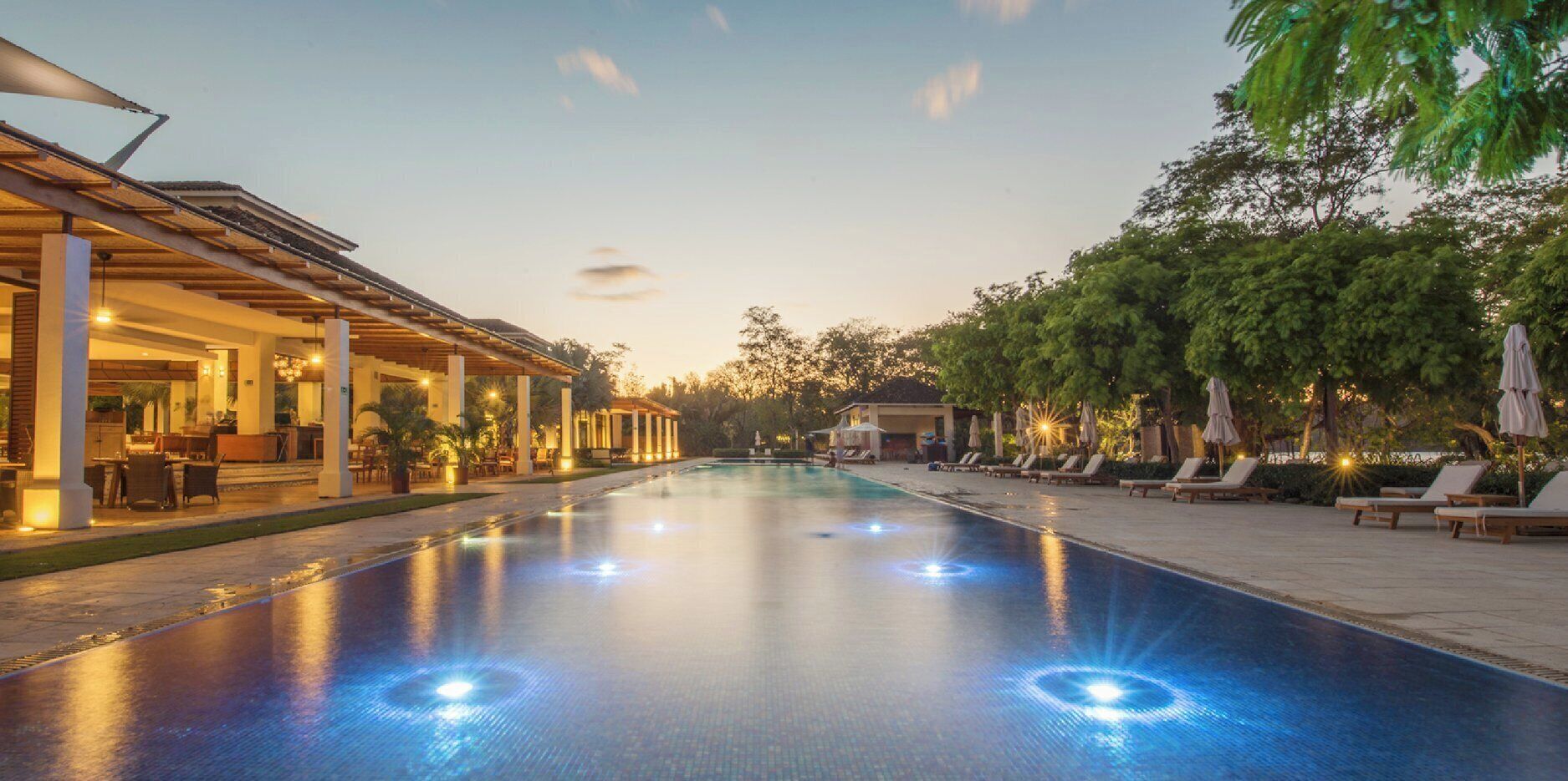 A swimming pool with a covered area and lounge chairs on one side, and a building with outdoor seating on the other.