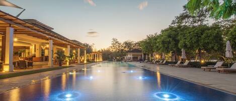 A swimming pool with a covered area and lounge chairs on one side, and a building with outdoor seating on the other.