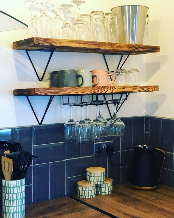 A kitchen with wooden shelves holding glassware and mugs, a coffee maker, and a patterned container on the counter. - Lusty glaze (Newquay)