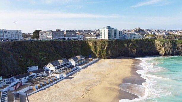 A coastal hotel with a sandy beach, a cliffside, and a view of the town in the background.