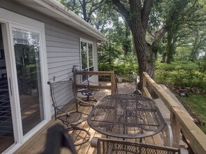 A covered patio with a table and chairs, a wooden deck, and a view of a wooded area.