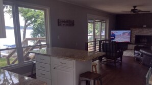 Una cocina con encimera de granito, armarios blancos y un taburete de bar. Hay un televisor en la sala de estar con chimenea.