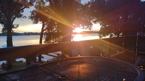 Un espacio circular con asientos al aire libre, una parrilla de metal, rodeado de árboles y con vista a un cuerpo de agua durante el atardecer.