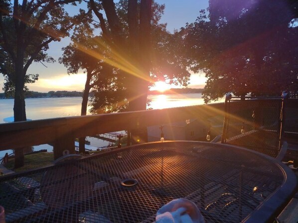 A circular outdoor seating area with a metal grill, surrounded by trees and overlooking a body of water during sunset.