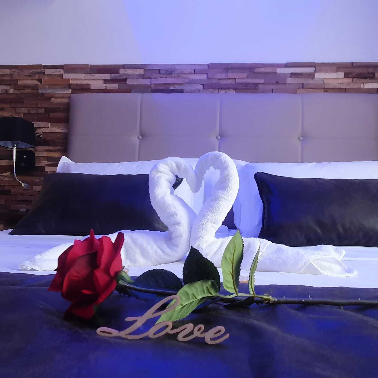 Ein Bett mit einem herzförmig gefalteten weißen Handtuch, einer roten Rose und einem 'Love'-Schild auf einer blauen Bettdecke.