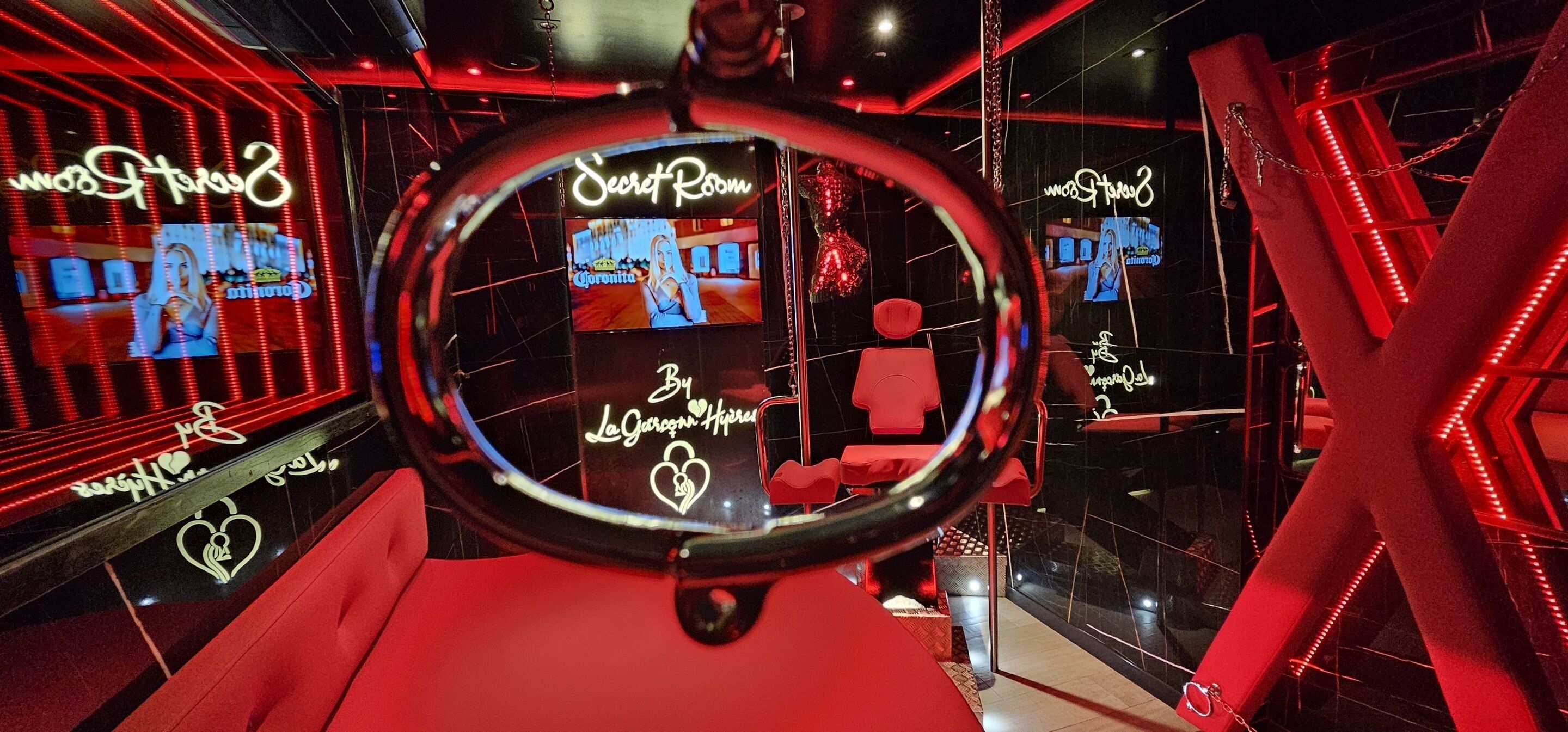 A room with a circular mirror, red seating, and neon signs reading "Secret Room" and "By Le Gourgonnier."
