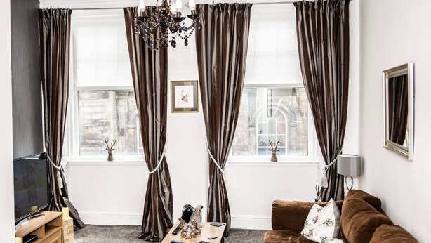 A living room with a brown sofa, a wooden coffee table, and a chandelier. There are two windows with curtains, a framed picture on the wall, and a television on the left. - Rose Apartment 2-bed Apt Town Centre Ayr (Ayr)