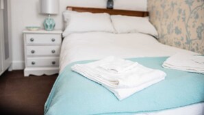 A neatly made bed with a light blue bedspread and white towels, a wooden headboard, a white nightstand with drawers, a lamp, and a vase with flowers. - Rose Apartment 2-bed Apt Town Centre Ayr (Ayr)