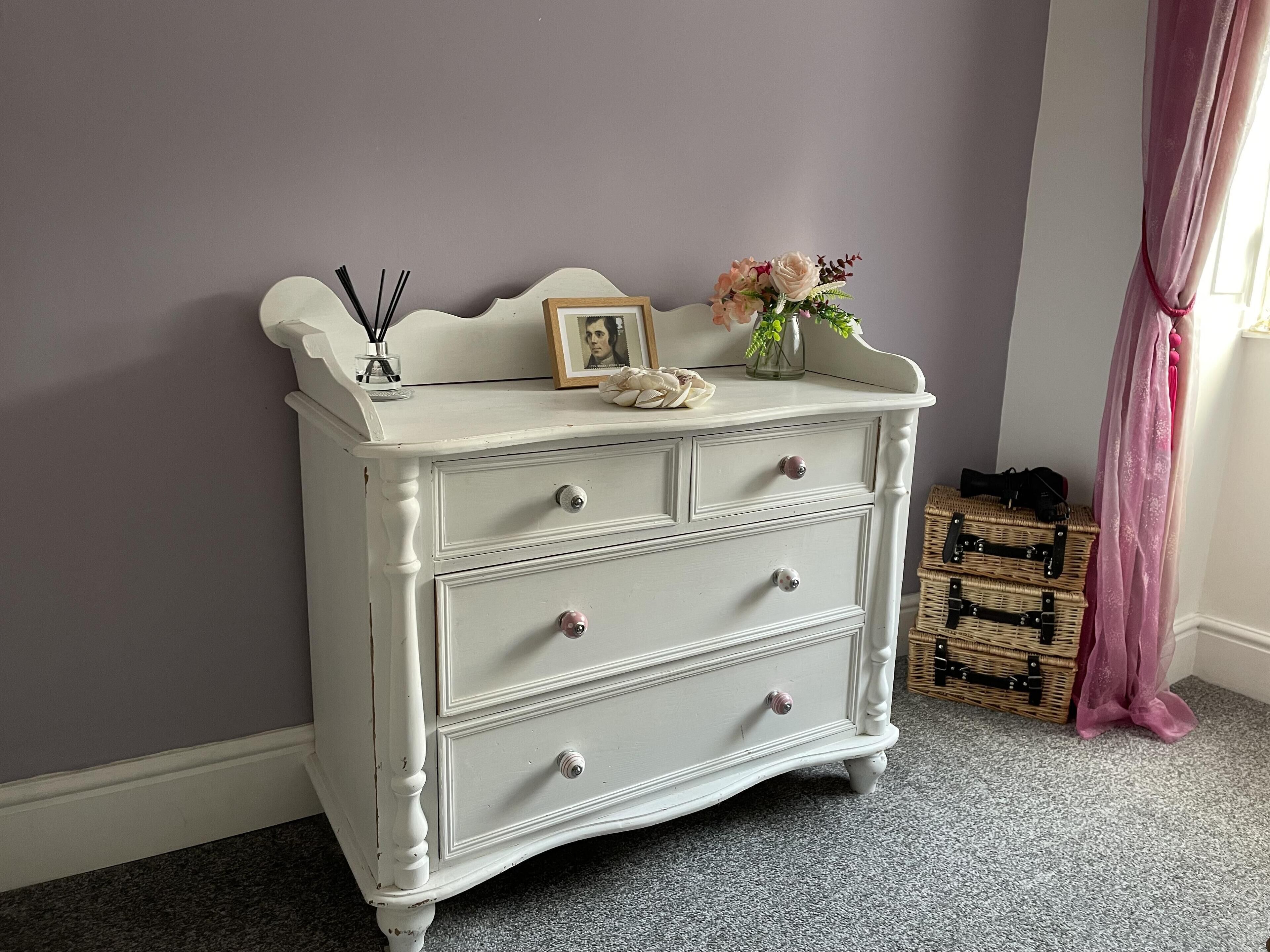 Une commode blanche à trois tiroirs, un cadre photo et un vase avec des fleurs posé sur le dessus.