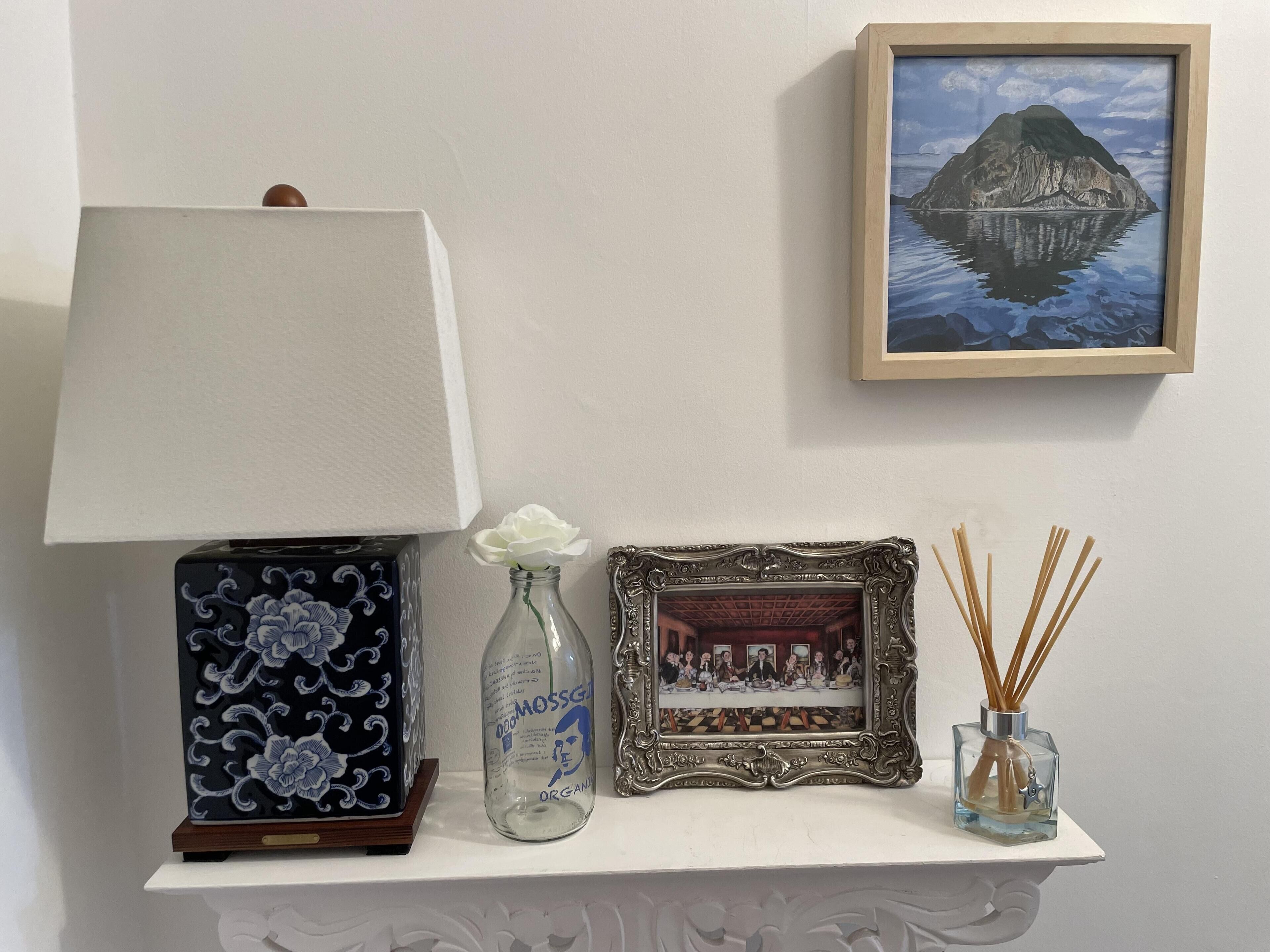 A table with a lamp, a framed picture, a bottle, and reed diffusers.