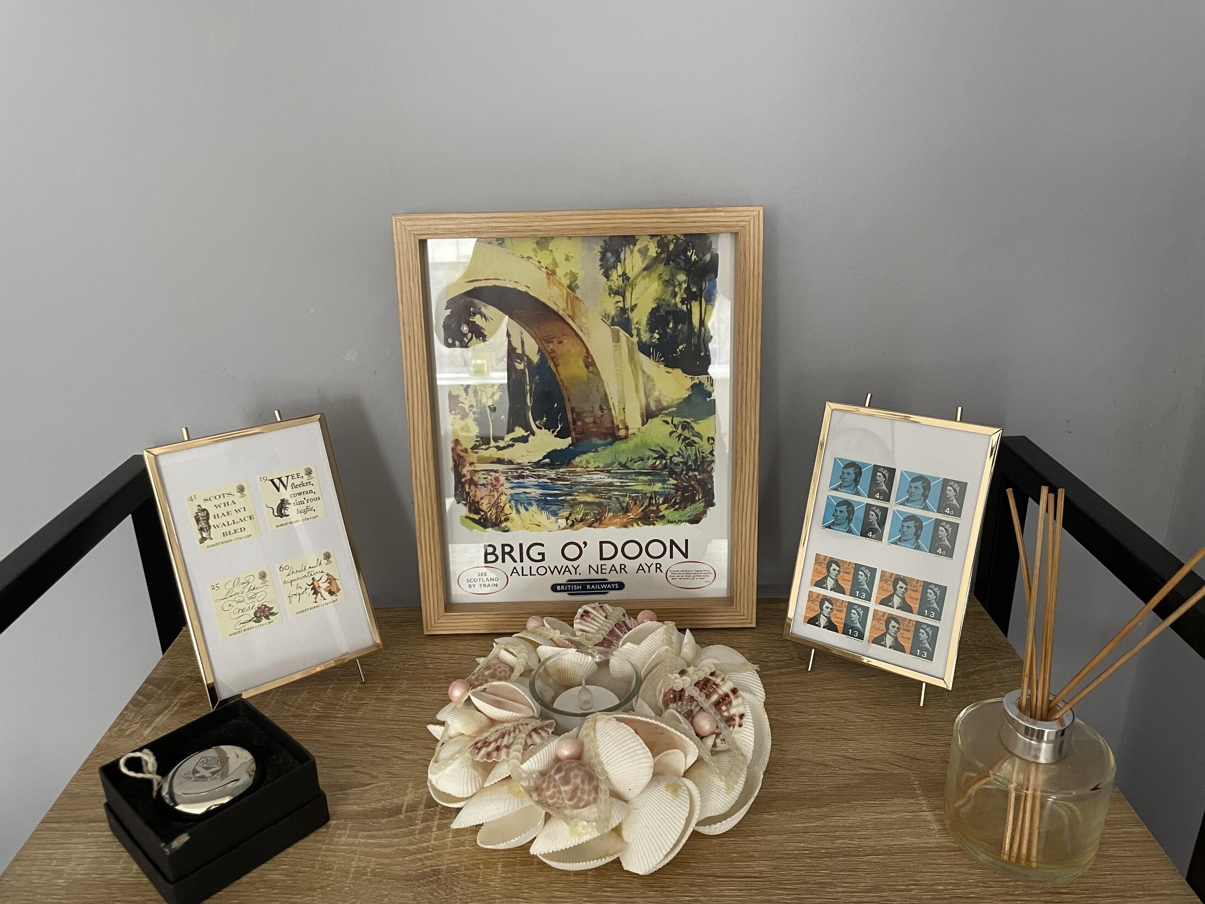 A framed artwork of Brig O'Doon, a vintage railway poster, surrounded by decorative shells and a small candle holder.
