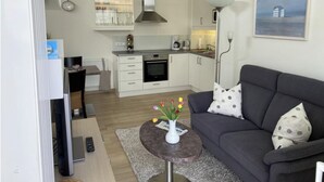 A modern living room with a grey sofa, a coffee table with a vase of flowers, and a kitchen area with white cabinets and a stove. - Apartment 1 city center, central, close to the beach and quiet (Norderney)