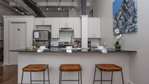 A modern kitchen with white cabinets, a stainless steel refrigerator, and a marble countertop. There are three wooden stools and a painting on the wall. - Downtown Fort Worth Luxury Loft in Sundance Square (Fort Worth)