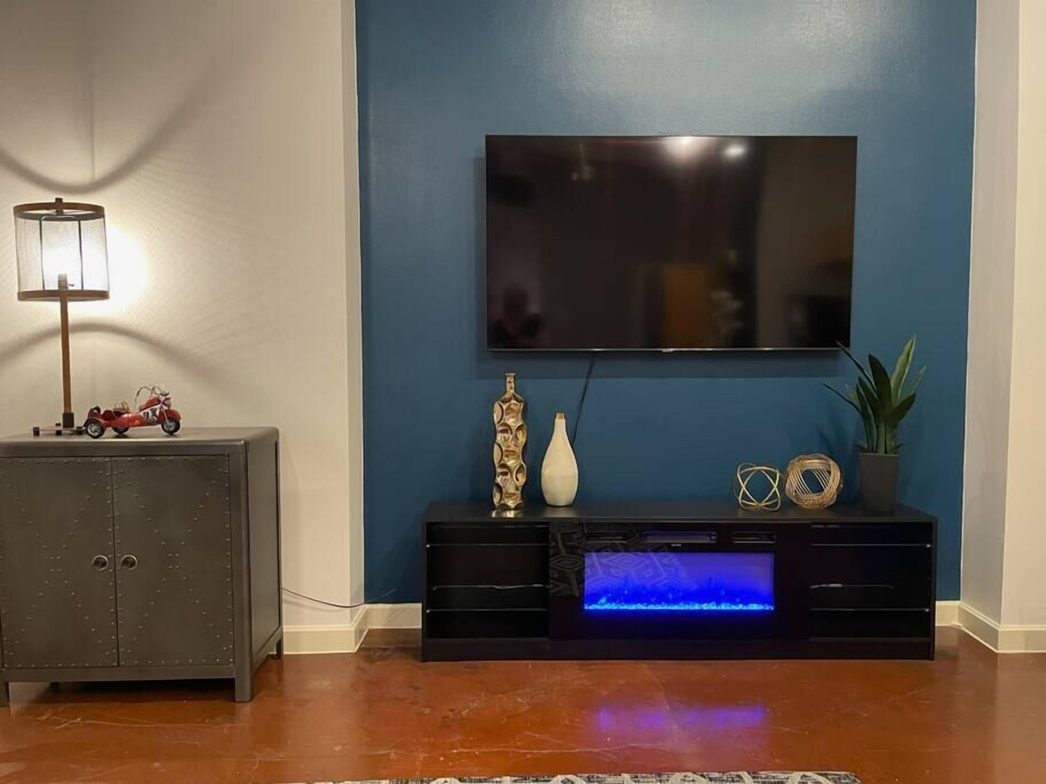 Sala de estar com televisão de tela plana presa a parede azul, móvel multimídia escuro com luz azul, pequeno armário com abajur e objetos decorativos.