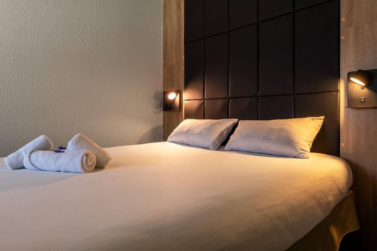 Ein ordentlich bezogenes Bett mit Kissen und einem zusammengerollten Handtuch, ein Kopfteil mit einer Leuchte und eine Wandlampe.