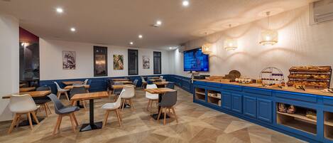 Un café moderno con mesas y sillas de madera, una barra azul con exhibición de alimentos y obras de arte en las paredes.
