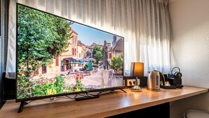 A flat-screen TV on a wooden desk with a remote, a kettle, and a lamp. - Hôtel Le Cyrano (Bergerac)