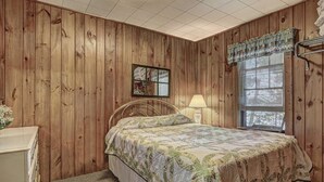 Une chambre avec des murs recouverts de panneaux de bois, un lit simple avec une couvre-lit à motifs floraux, une lampe de chevet, une fenêtre avec des stores et un petit miroir accroché au mur.