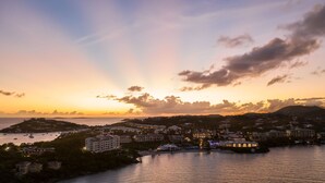 Une ville côtière au coucher du soleil, avec des bâtiments le long du front de mer, une marina remplie de bateaux et une colline en arrière-plan.