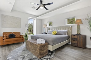 A modern bedroom with a large bed, a brown leather sofa, a wicker ottoman, and a ceiling fan.