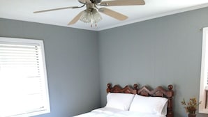 Un dormitorio con cama, cabecero de madera, ventilador de techo, mesita de noche y ventana con persianas.
