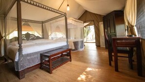 A four-poster canopy bed with a wooden bench in front, a wooden desk, and a view of the outdoors through the tent's entrance. - Lorian Safari Camp (Maasai Mara)