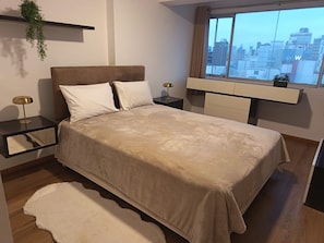 A modern bedroom with a bed, bedside tables, a dresser, a window with a city view, and a hanging plant.