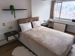 A modern bedroom with a large bed, bedside tables, a hanging plant, and a city view through the window.