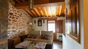 A cozy living room with a stone wall, wooden ceiling beams, a sofa with cushions, a table with a magazine, and a window with blinds.