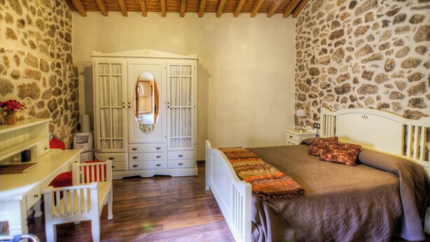 A bedroom with a stone wall, wooden ceiling beams, a white wardrobe, a bed with a brown blanket, and a small desk with a chair.