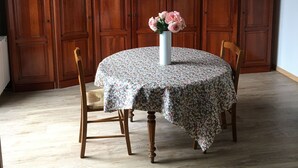 A dining area with a round table covered by a floral tablecloth, two wooden chairs, and a vase with flowers. The room has wooden paneling on the walls and a wooden floor. - Attractive 32 M2 studio with 12 M2 private courtyard (Dinard)