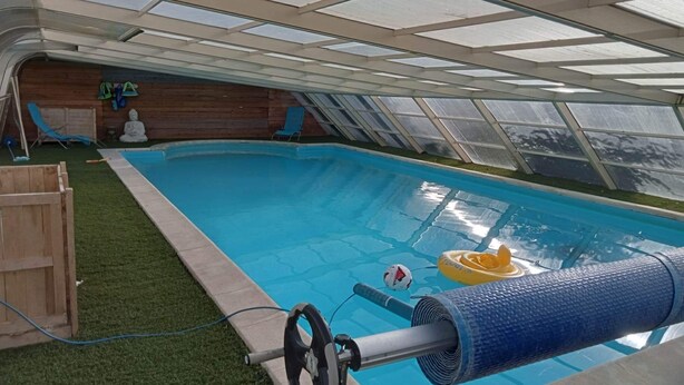 Una piscina con techo transparente, rodeada de césped artificial y terraza de madera.