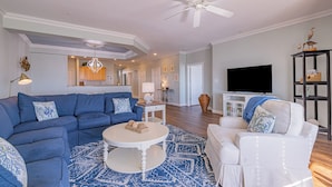 A living room with a blue sectional sofa, a white armchair, a coffee table, and a TV. - Book Beach Haven 213D | Oceanfront Sandbridge Rental (Virginia Beach)