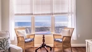 A room with a large window, wicker chairs, a round table, and a view of the ocean. - Book Beach Haven 213D | Oceanfront Sandbridge Rental (Virginia Beach)