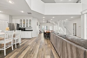 A modern kitchen with white cabinetry, a stainless steel refrigerator, and a dining area with a long table and chairs. - Book Spinnaker | Semi-Oceanfront Sandbridge Vacation Rental (Virginia Beach)