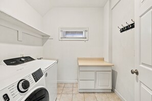 A laundry room with a front-loading washer and dryer, a small window, and a door with a coat rack. - Book Spinnaker | Semi-Oceanfront Sandbridge Vacation Rental (Virginia Beach)