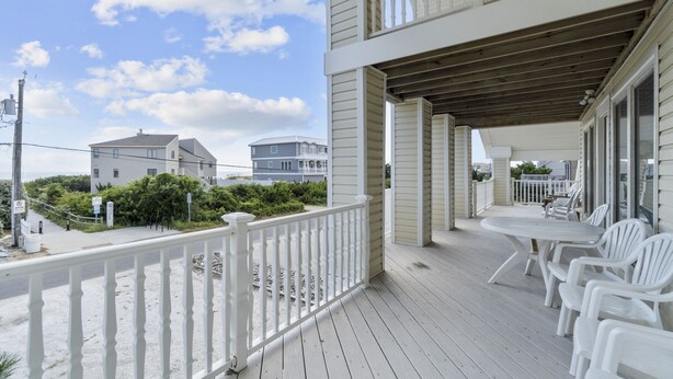 A wooden deck with white railings, a table with chairs, and a view of the ocean and other buildings. - Book Spinnaker | Semi-Oceanfront Sandbridge Vacation Rental (Virginia Beach)