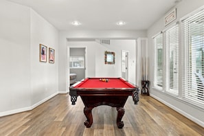 A pool table in a room with hardwood flooring, white walls, and a window with blinds. - Book Spinnaker | Semi-Oceanfront Sandbridge Vacation Rental (Virginia Beach)