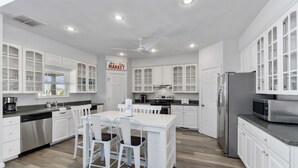 A modern kitchen with white cabinetry, a central island, and a dining area. - Book Spinnaker | Semi-Oceanfront Sandbridge Vacation Rental (Virginia Beach)