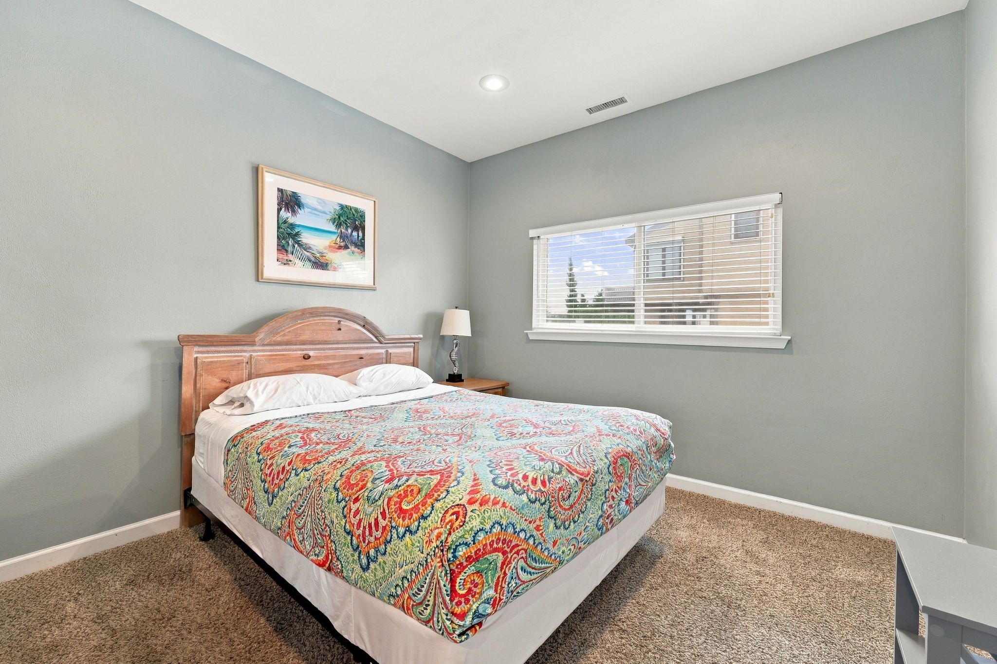 A bedroom with a bed, a nightstand, a lamp, a window with blinds, and a picture on the wall.