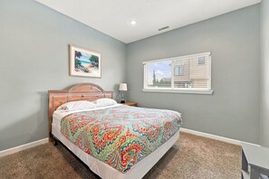 A bedroom with a bed, a nightstand, a lamp, a window with blinds, and a picture on the wall. - Book Spinnaker | Semi-Oceanfront Sandbridge Vacation Rental (Virginia Beach)