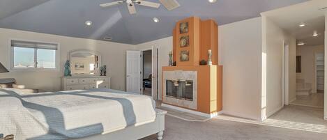 A bedroom with a bed, a ceiling fan, recessed lighting, a fireplace, and a hallway leading to another room.