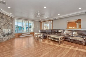 A spacious living room with a stone fireplace, large windows, a brown leather sectional sofa, and a ceiling fan. - Luxurious Beach Home with Indoor Pool Access! Steps from the Beach (Virginia Beach)