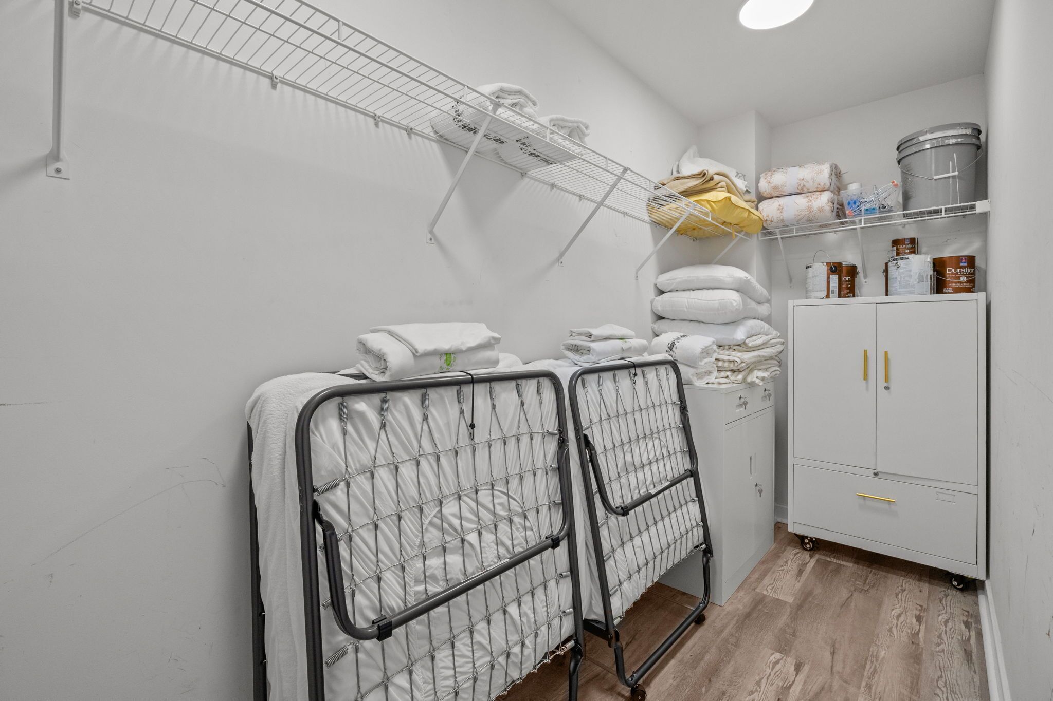 A room with two metal beds, a white cabinet with drawers, and a metal shelf with towels and supplies.