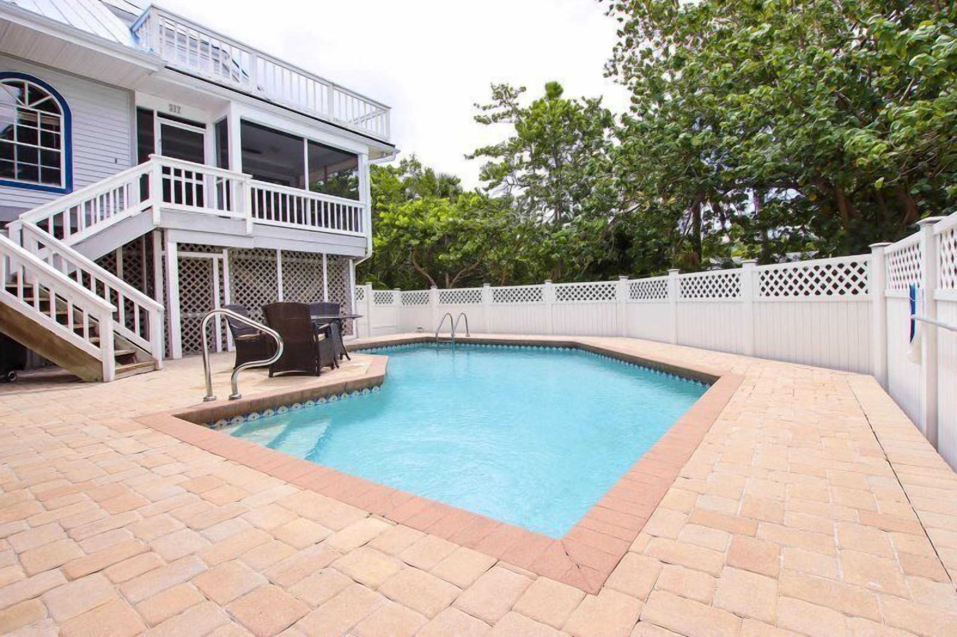 Une piscine avec une terrasse blanche, un espace de terrasse couvert et une clôture blanche.