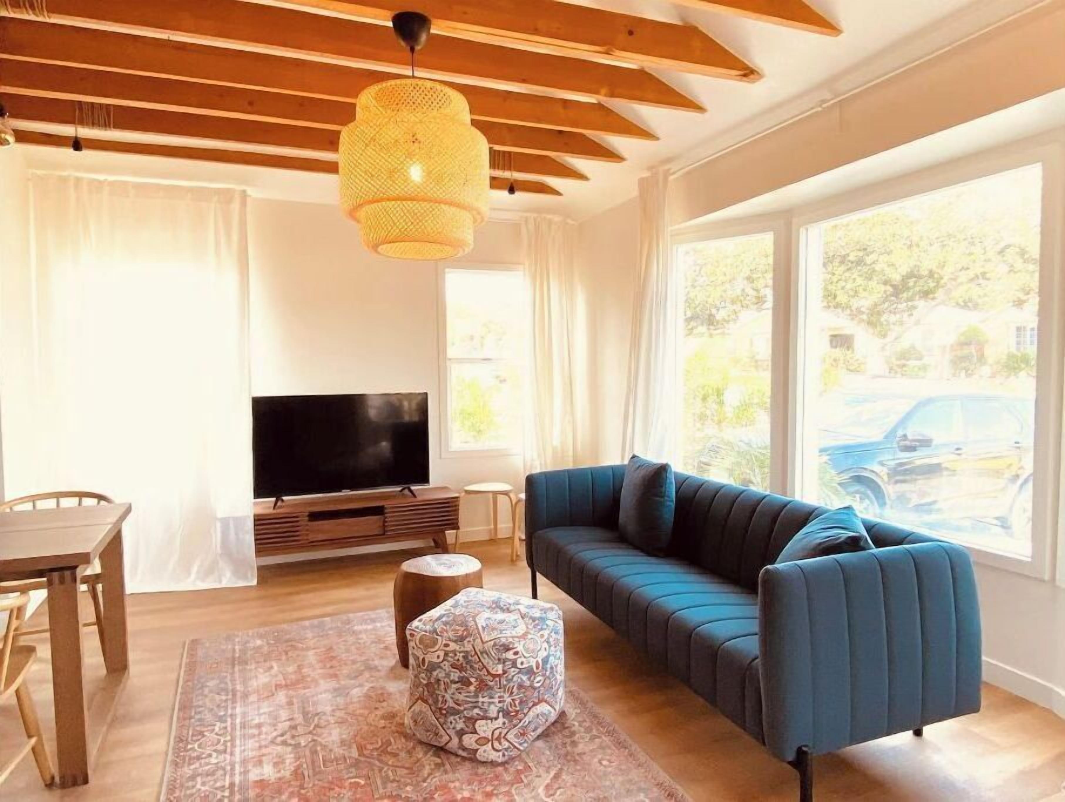 A modern living room with a large TV, a blue sofa, a patterned ottoman, and a wooden ceiling.