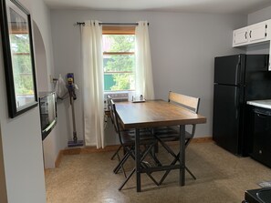 A compact kitchen with a wooden table, black chairs, a black refrigerator, and a window with white curtains.