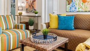 Un salon avec un canapé à rayures, un divan marron agrémenté de coussins jaunes et bleus, et une table basse en osier sur laquelle est posée une plante.
